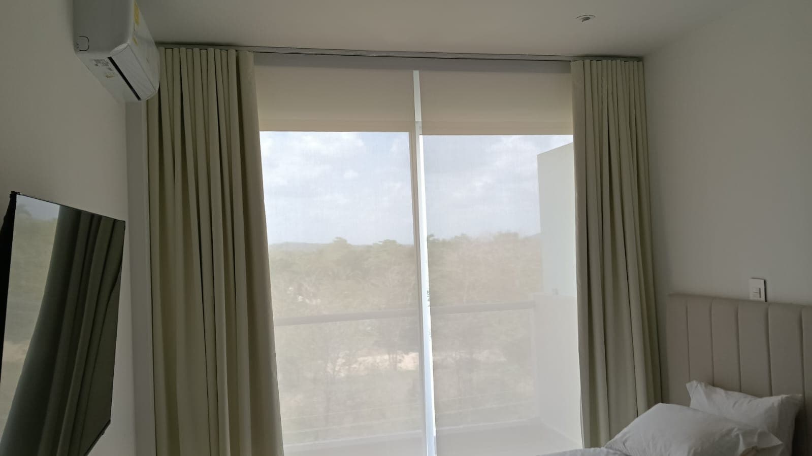 Cortinas pliegue cream con persiana solar screen desenrollada detrás en habitación con vista a vegetación