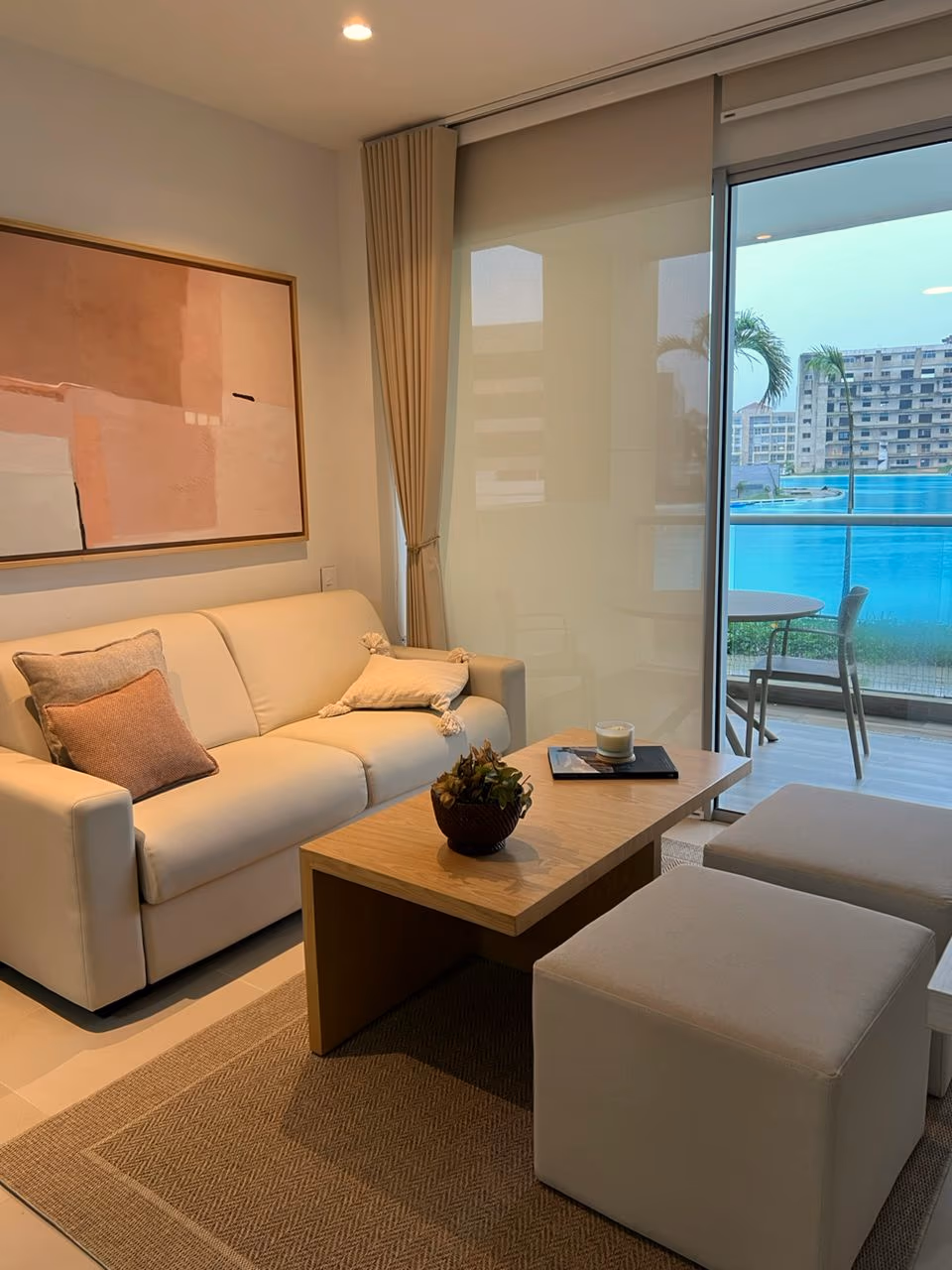 Sala pequeña con sofá crema, mesa de centro de madera, puffs y terraza con laguna