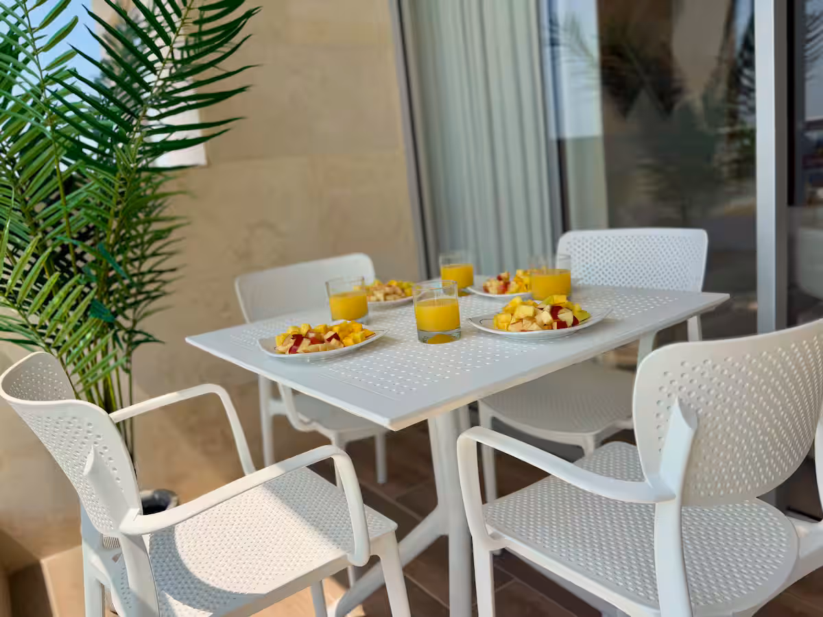 Terraza con mesa blanca, frutas, jugos y una palma